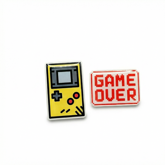 Two enamel pins: one resembling a vintage game console and the other 'GAME OVER' on a white background.